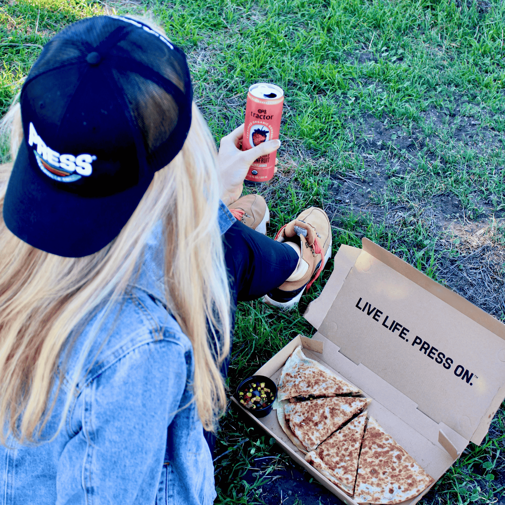 A young woman sitting on the grass with a Press quesadilla to-go.