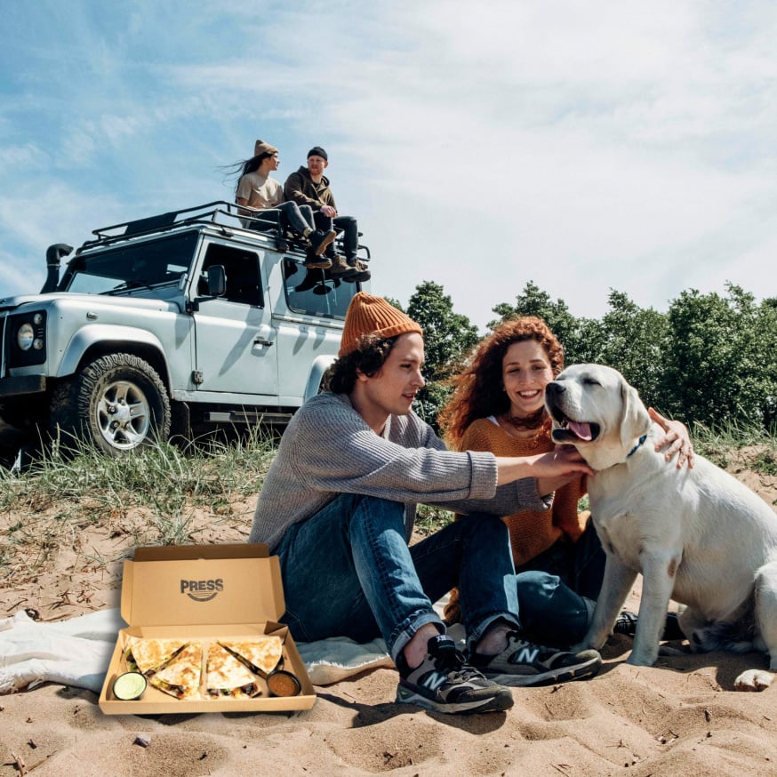 Two people petting a labador dog while sitting next to a Press to-go box.