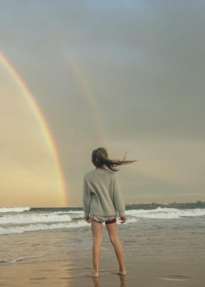 A young child standing on a beach staring at a rainbow.